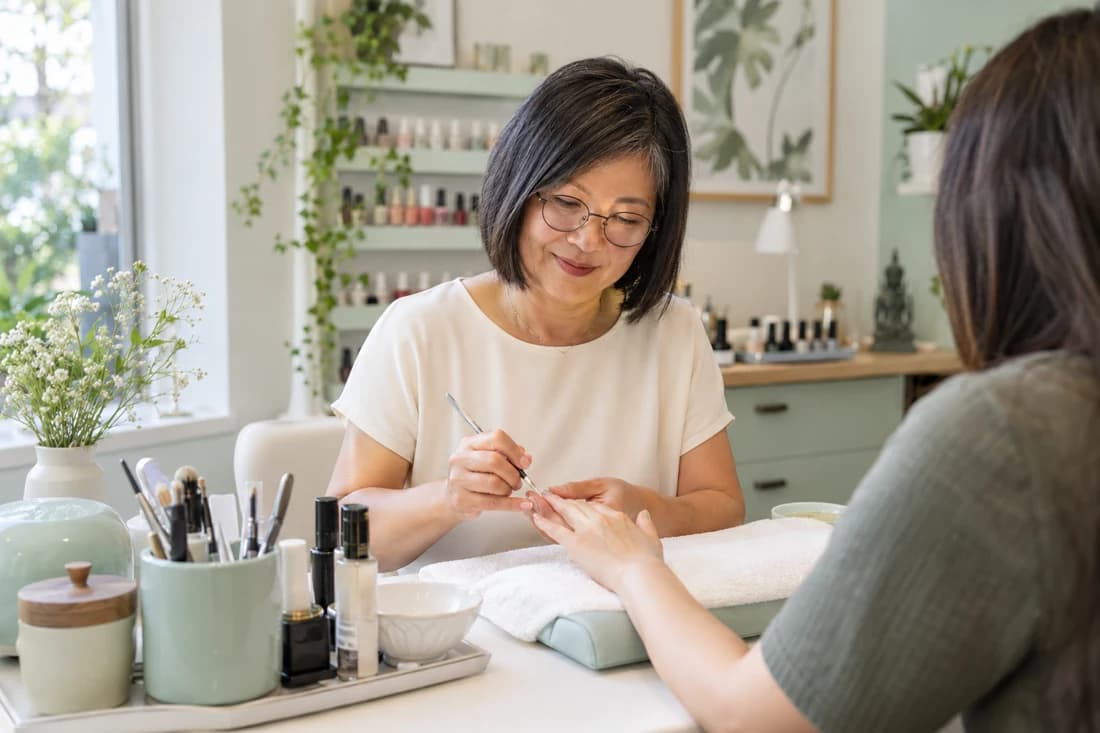 Een nagelstyliste met een bobkapsel en ronde bril verzorgt de nagels van haar klant